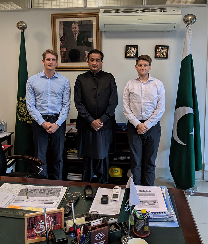 Three men standing behind a table. Official Pakistan flags are next to them